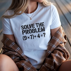 May include: White t-shirt with the text "SOLVE THE PROBLEM (9x7)+4=?" in bold black letters. The person is wearing a brown and tan plaid shirt over the white t-shirt. A gold necklace is visible.