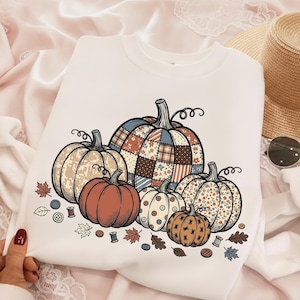 May include: A white crewneck sweatshirt featuring a graphic print of five pumpkins in various patterns, including plaid, floral, and solid orange. The design is accented with small scattered autumn leaves, sewing buttons, and thread spools.