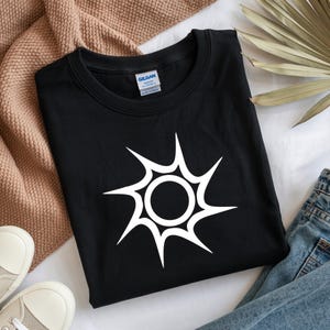 Puede incluir: Camiseta negra con un diseño blanco de explosión solar. El gráfico tiene un círculo central con líneas puntiagudas que irradian. La camiseta está doblada sobre una superficie neutra, con zapatillas blancas y vaqueros azules visibles.