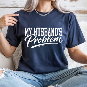 Könnte beinhalten: Marineblaues T-Shirt mit dem weißen Text "MY HUSBAND'S Problem". Das Shirt besteht aus einem weichen Material. Das Shirt ist ein Freizeitkleidungsstück.