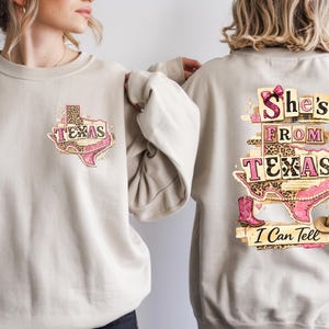 Może przedstawiać: Beżowa bluza z grafiką w stylu Teksasu. Prz&oacute;d przedstawia kontur Teksasu ze słowem "TEXAS" oraz r&oacute;żowym i lampartowym wzorem. Tył wyświetla "She's From Texas I Can Tell" z konturem Teksasu, kowbojskimi butami i kapeluszem kowbojskim.