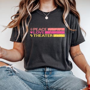 May include: Dark grey t-shirt with the words "PEACE LOVE THEATER" in pink, orange, and yellow. The word "PEACE" has a peace symbol, "LOVE" has a heart, and "THEATER" has a theatre mask.