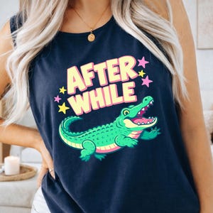 May include: Dark blue vest top with a cartoon alligator design and the text "AFTER WHILE". The alligator is green with pink details, and the text is in yellow and pink. The design includes yellow and pink stars.