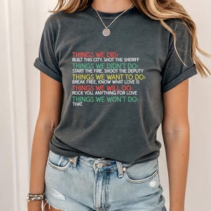 以下が含まれることがあります： カラフルなテキストが入った濃いグレーのTシャツ。テキストは、「THINGS WE DID」、「THINGS WE DIDN'T DO」、「THINGS WE WANT TO DO」などの見出しの下にフレーズをリストしています。このTシャツは、ブルージーンズとシルバーのネックレスを身に着けた人が着用しています。