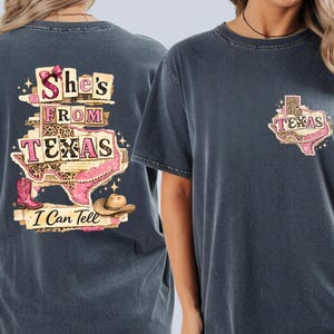 Może przedstawiać: Ciemnoszara koszulka z motywem Teksasu. Z przodu ma mały zarys Teksasu z nadrukiem w cętki i napisem "Texas". Z tyłu napis "She's From Texas I Can Tell" z zarysem Teksasu, kowbojskimi butami i kapeluszem kowbojskim.