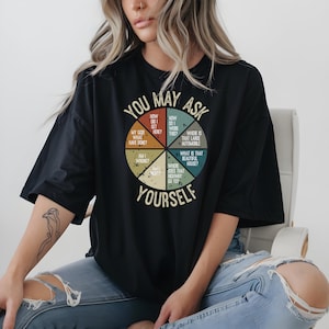 May include: Black t-shirt featuring a retro pie chart graphic with phrases such as "How do I get here?" and "Where is that large automobile?" The text "YOU MAY ASK YOURSELF" is also included.