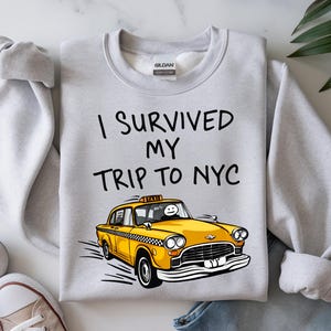 May include: A light gray sweatshirt with the text "I Survived My Trip To NYC" above a yellow taxi cab graphic. The taxi has a black and white checkered pattern and a smiling face in the driver's seat.