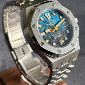 May include: A silver-coloured wristwatch with a blue and black patterned face. The watch features gold-coloured hands and markers, a date display, and a silver-coloured strap. The watch is displayed on a wooden stand.