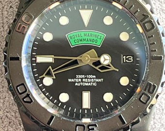 May include: Black wristwatch with a black dial. The watch face features the text "Royal Marines Commando" in a green rectangle. The watch has a black bezel with white markings and a black metal strap. The watch face also displays the date.