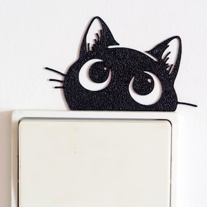 Puede incluir: Una cubierta de interruptor de luz con forma de gato negro con ojos y bigotes blancos, montada sobre un interruptor de luz blanco. La cabeza del gato tiene textura y parece estar hecha de un material mate. El fondo es una pared blanca lisa.