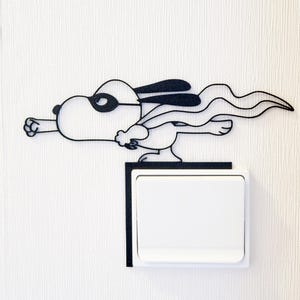 Può includere: Decorazione murale in metallo nero con Snoopy supereroe. Il design presenta Snoopy con maschera e mantello, che vola sopra un interruttore della luce bianco. L'opera d'arte è montata su una parete bianca strutturata.