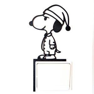 May include: Black metal Snoopy wall decor. Snoopy is wearing a nightcap and pajamas. The Snoopy figure is standing on a black rectangular shelf with a white square below it. The background is white.