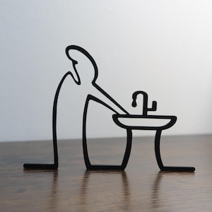 Könnte beinhalten: Schwarze Metallsculptur einer Person, die sich an einem Waschbecken die Hände wäscht. Die Skulptur ist minimalistisch gestaltet mit einer einfachen Silhouette.