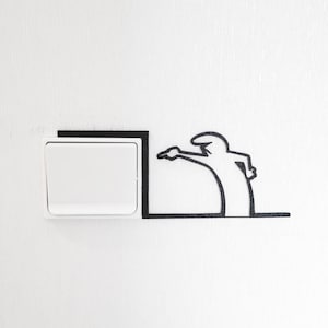 Può includere: Un interruttore della luce bianco con un contorno nero è su una parete bianca. Una silhouette nera di una persona sta puntando l'interruttore.
