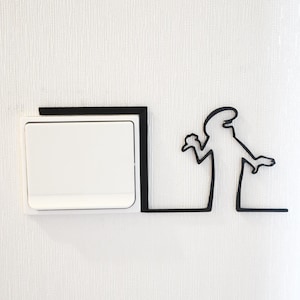 May include: A white light switch with a black outline is mounted on a white wall. A black silhouette of a person with outstretched arms is drawn on the wall next to the switch.