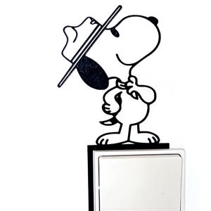 May include: Black and white Snoopy-themed light switch cover. The design features Snoopy wearing a hat and holding a stick, standing above a white light switch. The cover has a black border.