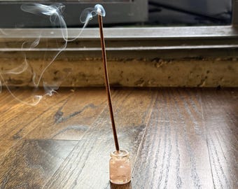 以下が含まれることがあります： 細い茶色の線香が、淡いピンク色の半透明で不規則な形の石の破片が入った小さな透明なガラス瓶に立てられています。線香の火がついた先端から白い煙が立ち上り、空中に漂っています。