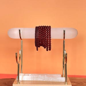 May include: A collection of deep red, spherical bead bracelets draped over a horizontal, translucent white cylindrical bar. The bar is supported by a gold-toned metal stand with a wooden base, featuring an additional white rectangular block.