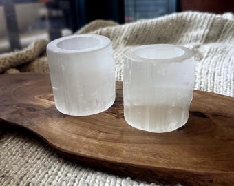 以下が含まれることがあります： 半透明の白いセレナイトクリスタルで作られた2つの円筒形キャンドルホルダー。各ホルダーは、滑らかで磨かれた上部の縁と、外壁に沿った自然な繊維状の垂直テクスチャが特徴で、鉱物の結晶構造を示しています。