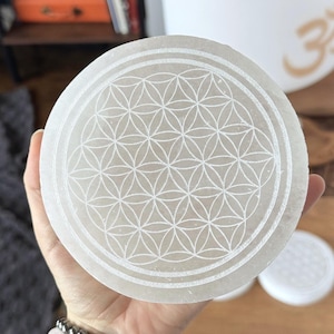 May include: A circular white selenite crystal charging plate featuring an etched flower of life geometric pattern on its surface. The translucent stone has a smooth, flat texture with a repeating series of intersecting circles forming a symmetrical design.