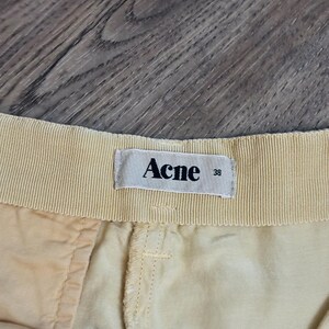 以下が含まれることがあります： ベージュのショートパンツのウエストバンドのクローズアップ。ラベルには「Acne」とサイズ「38」と記載されています。ショートパンツは明るい色の生地で作られており、縫い目が見えます。