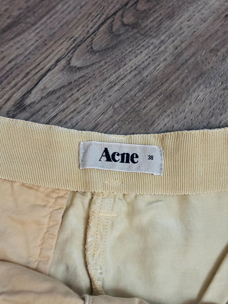 以下が含まれることがあります： ベージュのショートパンツのウエストバンドのクローズアップ。ラベルには「Acne」とサイズ「38」と記載されています。ショートパンツは明るい色の生地で作られており、縫い目が見えます。