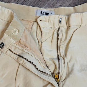 以下が含まれることがあります： ベージュ色のパンツのクローズアップ。ジッパーとボタンの留め具が見えます。「Acne」のブランド名がタグにプリントされています。パンツはプリーツのデザインで、軽くてシルキーな質感です。