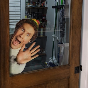 Puede incluir: Una puerta de madera marrón con un panel de vidrio. Un recorte de Buddy el Elfo de la película Elf está mirando a través del panel de vidrio. El panel de vidrio está detrás de un estante con varios artículos.