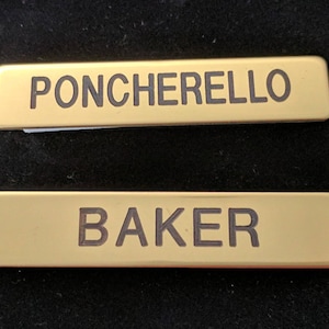 Puede incluir: Dos etiquetas de nombre doradas con letras negras. La etiqueta superior dice "PONCHERELLO" y la etiqueta inferior dice "BAKER".