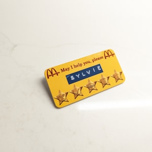 May include: Yellow McDonald's employee name tag with the text "May I help you, please" and the name "SYLVIE" in a blue rectangle. Five gold stars are displayed below the name.