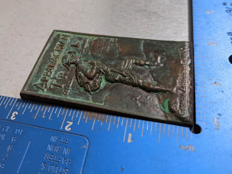 May include: A green metal plaque with the text "Appalachian Trail" and "The Georgia Appalachian Trail Club" embossed on it. The plaque features a raised image of a hiker carrying a backpack.