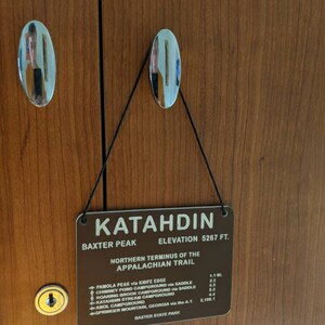 Puede incluir: Un letrero marr&oacute;n con el texto "Katahdin" en letras doradas. El letrero tambi&eacute;n incluye el texto "Baxter Peak, Elevation 5267 ft." y "Northern Terminus of the Appalachian Trail". El letrero enumera varias rutas de senderismo y distancias en millas.