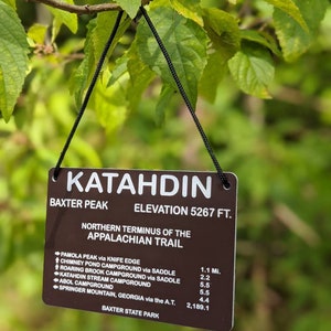 Puede incluir: Un letrero marr&oacute;n con texto blanco que dice "Katahdin Baxter Peak Elevation 5267 ft. Northern Terminus of the Appalachian Trail" y enumera las distancias a varios puntos a lo largo del sendero de los Apalaches, incluyendo Pamola Peak, Chimney Pond Campground, Roaring Brook Campground, Katahdin Stream Campground, Abol Campground y Springer Mountain, Georgia. El letrero est&aacute; colgado de un cord&oacute;n negro.
