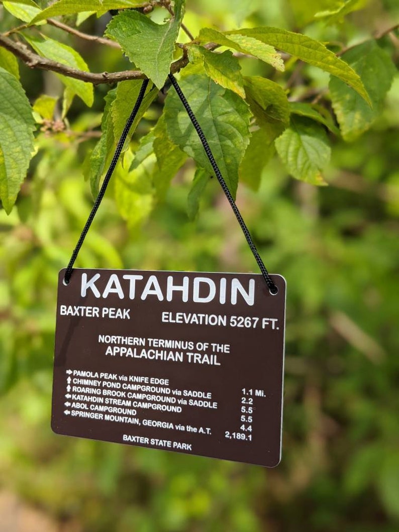 Puede incluir: Un letrero marr&oacute;n con texto blanco que dice "Katahdin Baxter Peak Elevation 5267 ft. Northern Terminus of the Appalachian Trail" y enumera las distancias a varios puntos a lo largo del sendero de los Apalaches, incluyendo Pamola Peak, Chimney Pond Campground, Roaring Brook Campground, Katahdin Stream Campground, Abol Campground y Springer Mountain, Georgia. El letrero est&aacute; colgado de un cord&oacute;n negro.