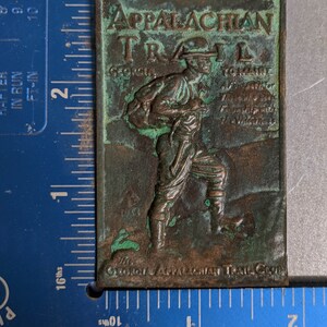 May include: A green and brown metal plaque with the words "Appalachian Trail" and "Georgia to Maine" embossed on it. The plaque features a hiker with a backpack and a walking stick.