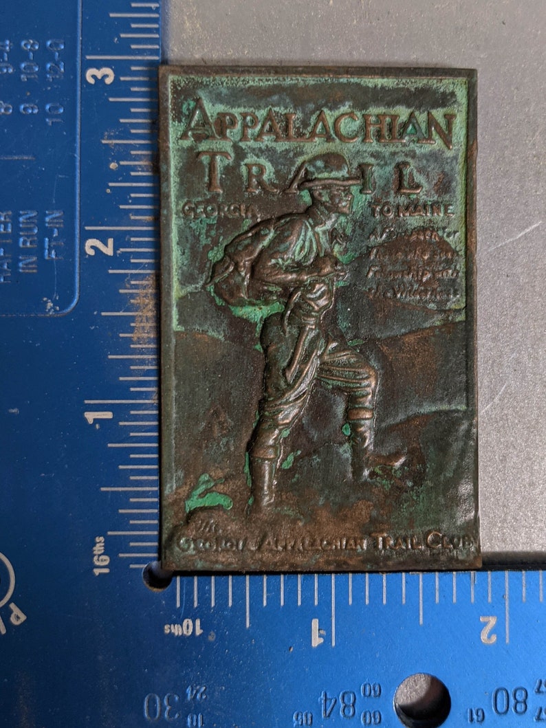 May include: A green and brown metal plaque with the words "Appalachian Trail" and "Georgia to Maine" embossed on it. The plaque features a hiker with a backpack and a walking stick.