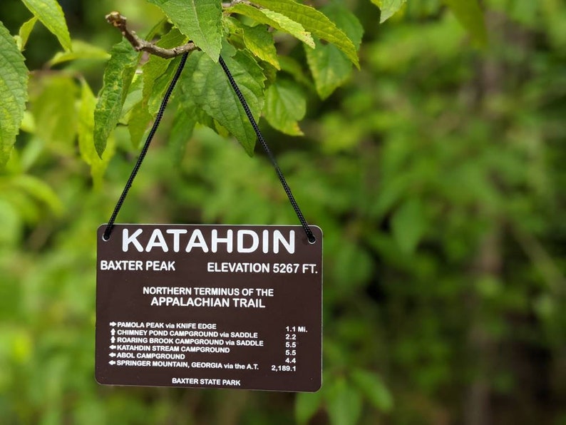 Puede incluir: Un letrero marr&oacute;n con texto blanco cuelga de una rama de &aacute;rbol. El letrero dice "Katahdin, Baxter Peak, Elevation 5267 ft. Northern Terminus of the Appalachian Trail". El letrero enumera las distancias a varios puntos a lo largo del sendero de los Apalaches, incluyendo Pamola Peak, Chimney Pond Campground, Roaring Brook Campground, Katahdin Stream Campground, Abol Campground y Springer Mountain, Georgia.