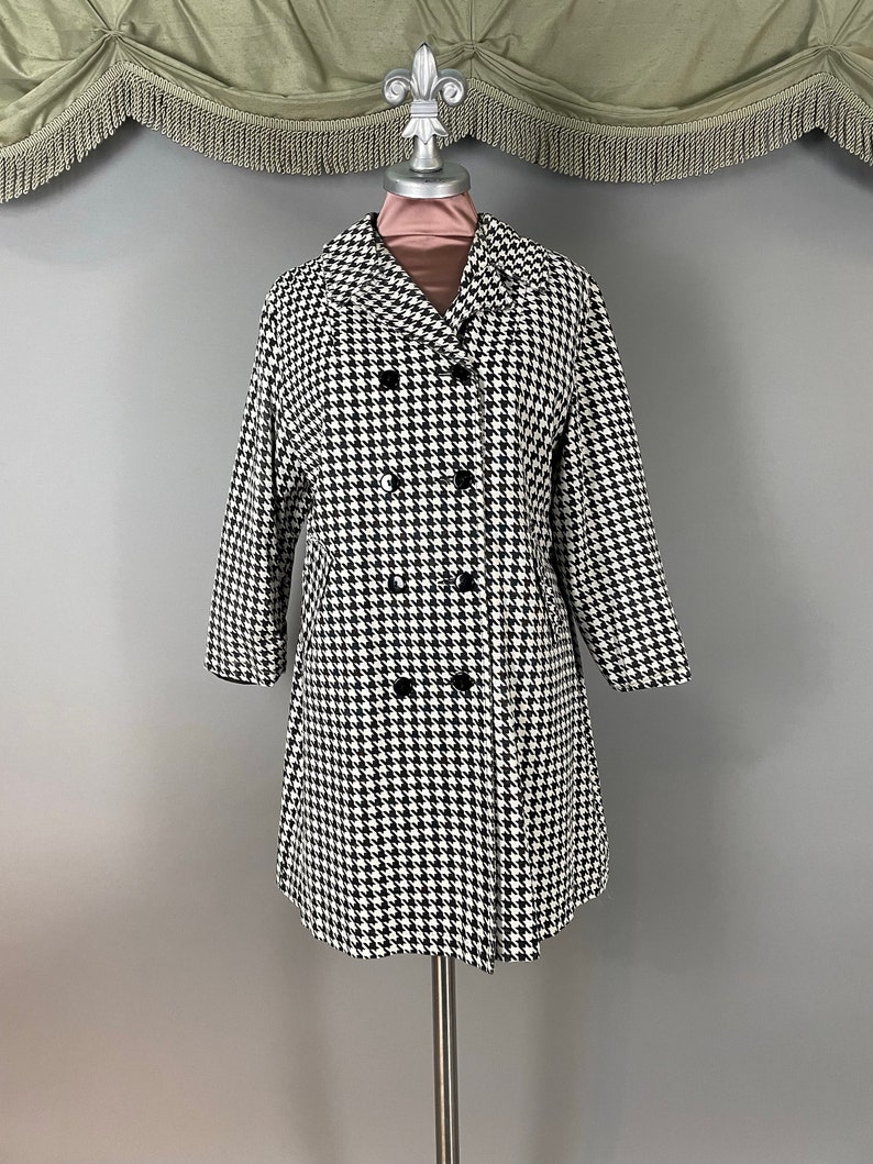 May include: A black and white houndstooth pattern coat with a double-breasted design. The coat has a classic collar, long sleeves, and black buttons. The coat is displayed on a mannequin.
