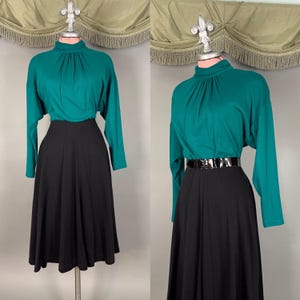 May include: A teal long-sleeve mock neck top with gathered detailing at the neckline paired with a black midi skirt. The outfit is displayed on a mannequin, with the right view showing a black patent leather belt cinched at the waist.
