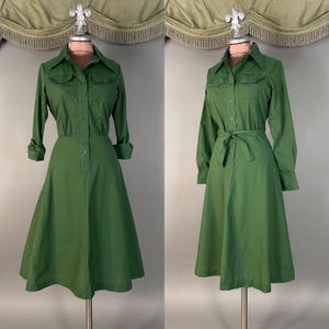 May include: A solid green midi dress featuring a collared neckline, a button-down front, and two chest pockets. The long sleeves have buttoned cuffs, and the waist includes a matching fabric tie belt that cinches the skirt into a flared silhouette.
