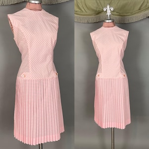 May include: A vintage pink dress with a checkered bodice and pleated skirt. The sleeveless dress features a round neckline and two decorative buttons. The dress is displayed on a mannequin.