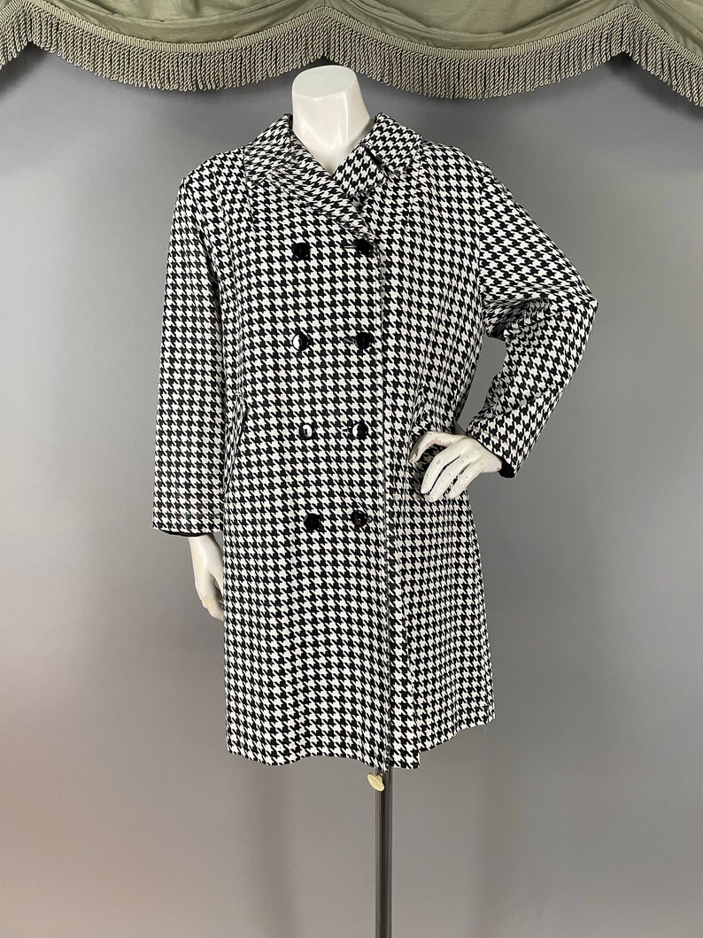 May include: A black and white houndstooth pattern coat with a double-breasted design. The coat has black buttons and a black trim on the collar and cuffs. The coat is displayed on a mannequin.
