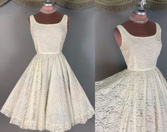May include: A sleeveless cream-coloured lace dress featuring a scoop neckline and a fitted bodice that transitions into a full, flared skirt. The intricate floral lace pattern covers the entire garment, which is displayed on a mannequin.