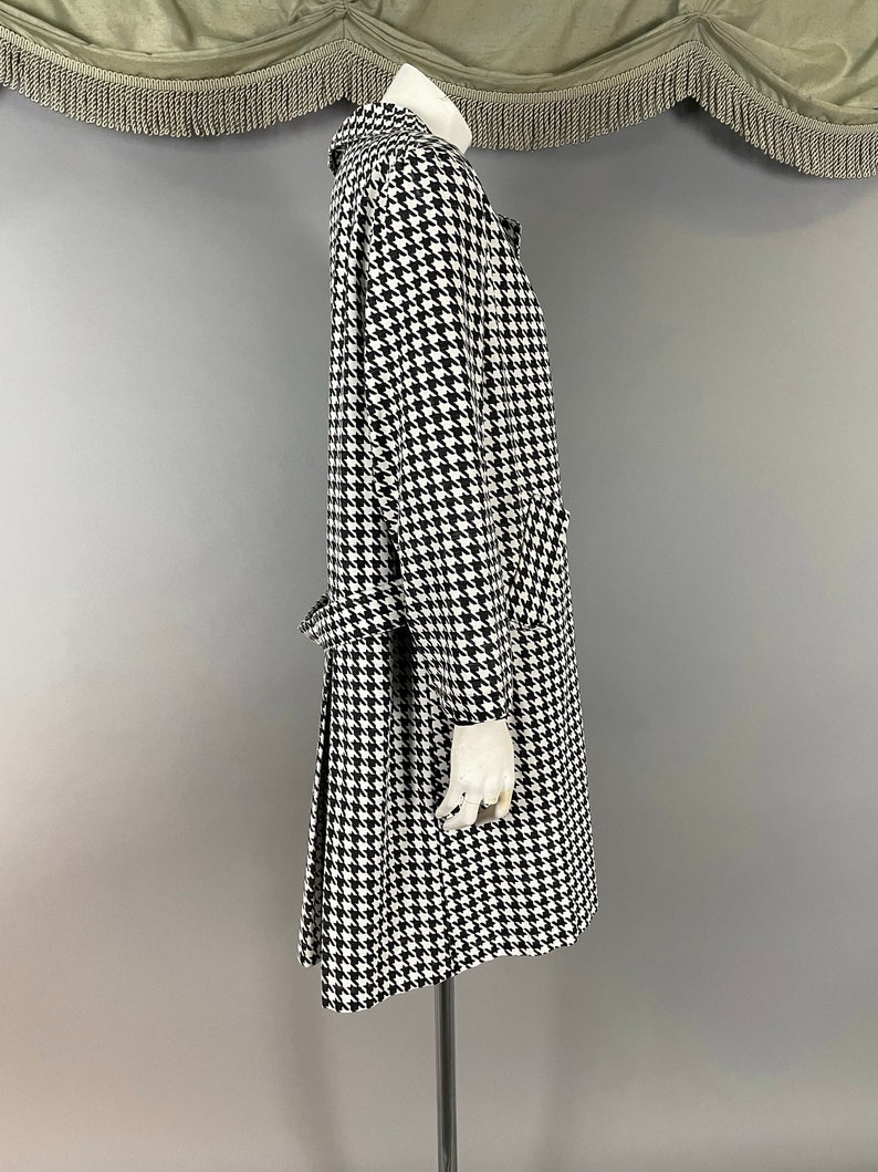 May include: A black and white houndstooth pattern coat with a collar, long sleeves, and a belt. The coat is displayed on a mannequin. The coat has a classic design and appears to be made of a medium-weight fabric.
