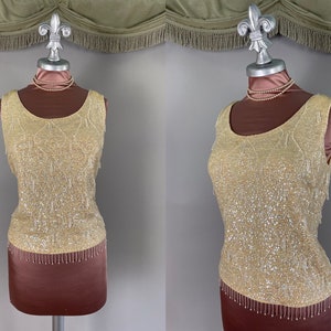 May include: A vintage, sleeveless, cream-colored top with intricate sequin and bead embellishments. The top features a scoop neckline and a fringe hemline. The garment is displayed on a mannequin with a pearl necklace.