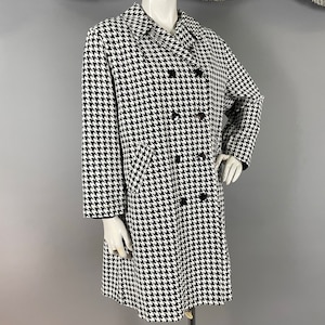 May include: A black and white houndstooth pattern coat on a mannequin. The double-breasted coat has black buttons and a notched collar. The coat is knee-length and has long sleeves. The mannequin is on a metal stand against a grey background.
