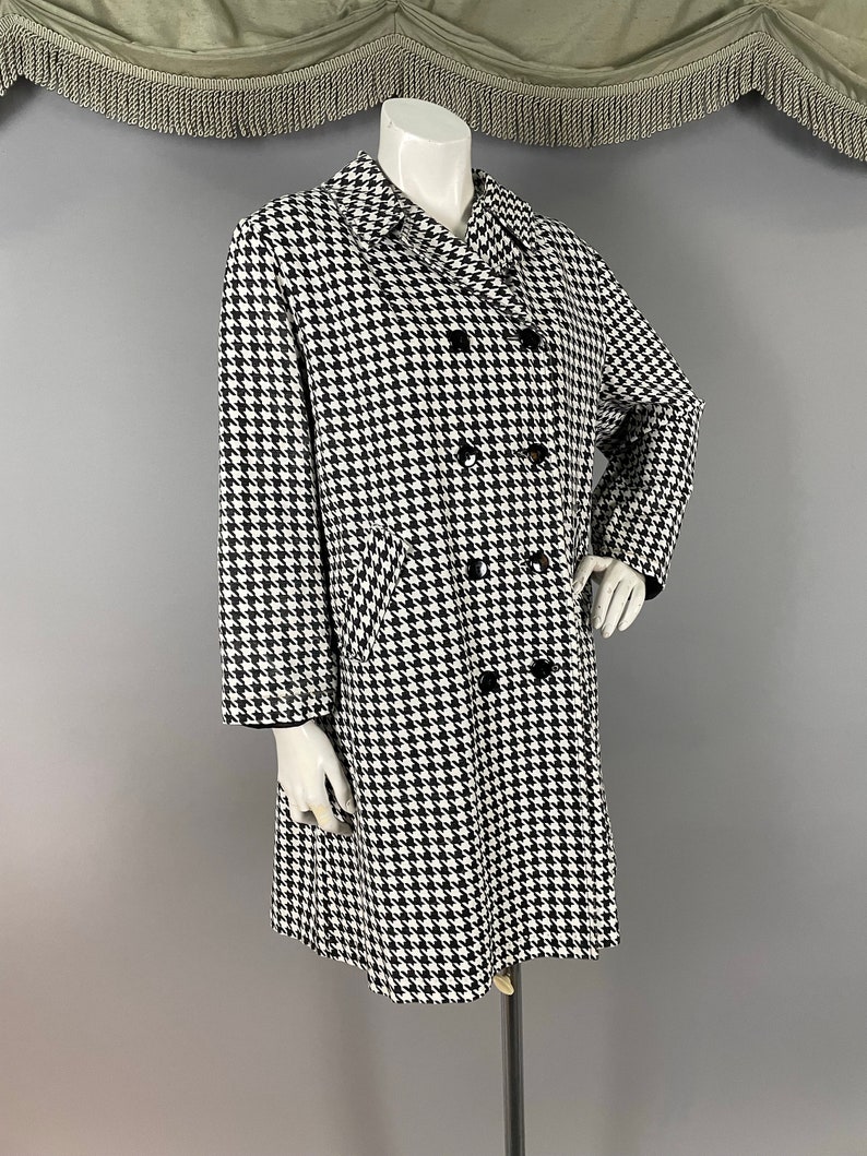 May include: A black and white houndstooth pattern coat on a mannequin. The double-breasted coat has black buttons and a notched collar. The coat is knee-length and has long sleeves. The mannequin is on a metal stand against a grey background.