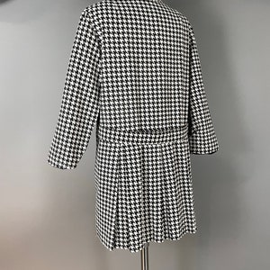 May include: A black and white houndstooth pattern coat with a pleated skirt. The coat is displayed on a mannequin. The coat has long sleeves and a belt. The coat is approximately 102 cm long.