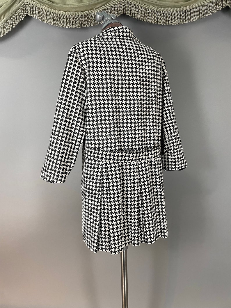 May include: A black and white houndstooth pattern coat with a pleated skirt. The coat is displayed on a mannequin. The coat has long sleeves and a belt. The coat is approximately 102 cm long.