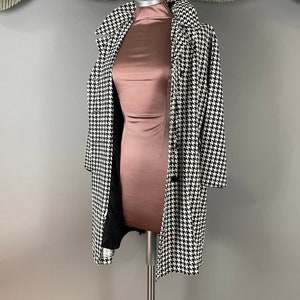 May include: A black and white houndstooth coat on a mannequin. The coat features a classic design with a notched collar and button closure. The mannequin is wearing a rose-coloured dress. The background is a neutral grey.